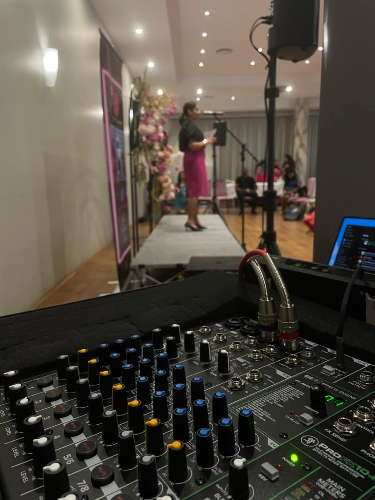 Studio AV's mixing desk at a smaller corporate conference, with a speaker at the lectern reading from notes against a floral backdrop